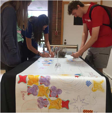 Group of students gathering at a table for the Election Day Creativity event. They draw and trace colorful designs on a long scroll of white craft paper.