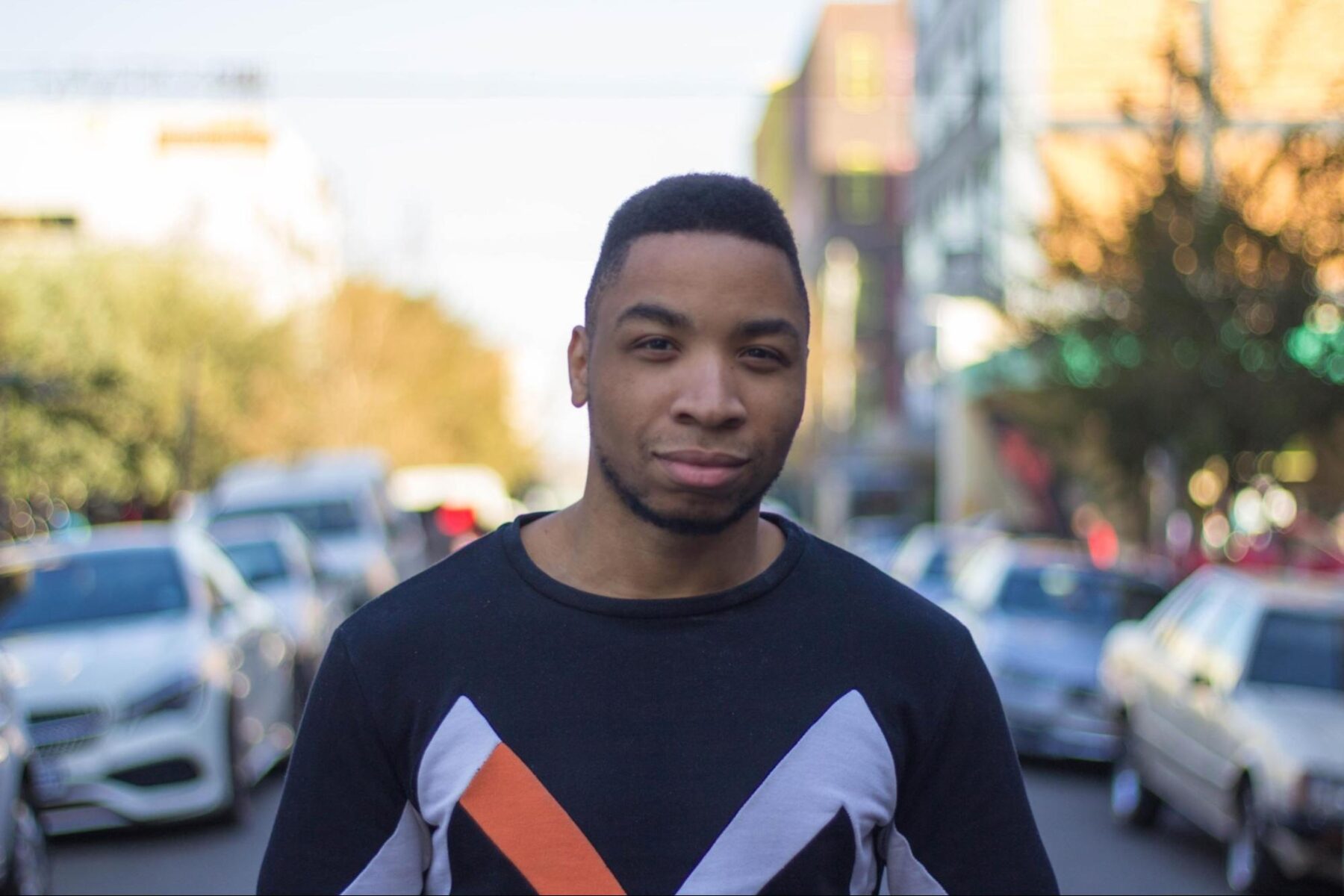 A man stands on a sunny city street, facing the camera. He wears a black sweater with lavender and orange accents.