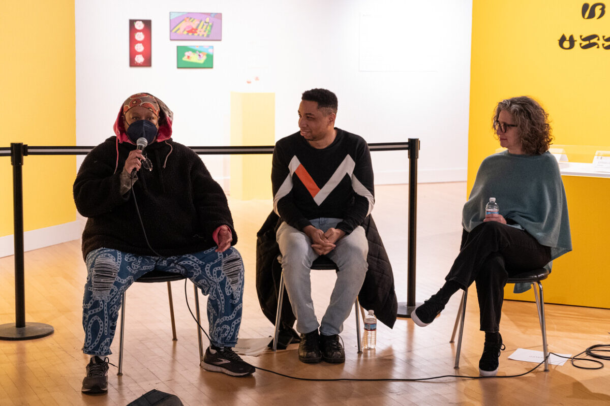 Sheila Gaskins, Levester Williams, and Lisa Freiman sit together for a discussion during Williams' exhibition opening. Gaskins at left holds a microphone and is speaking to an audience.
