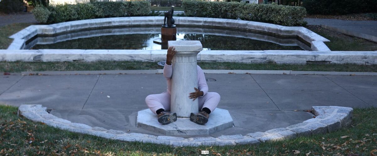 A barefoot woman dressed in white is seated on the ground behind a pillar. She is hugging the pillar, which is located in an outdoor public space.