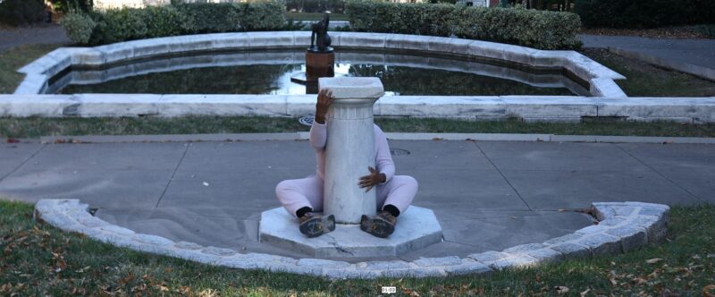 A barefoot woman dressed in white is seated on the ground behind a pillar. She is hugging the pillar, which is located in an outdoor public space.