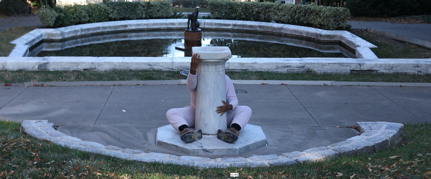 A barefoot woman dressed in white is seated on the ground behind a pillar. She is hugging the pillar, which is located in an outdoor public space.