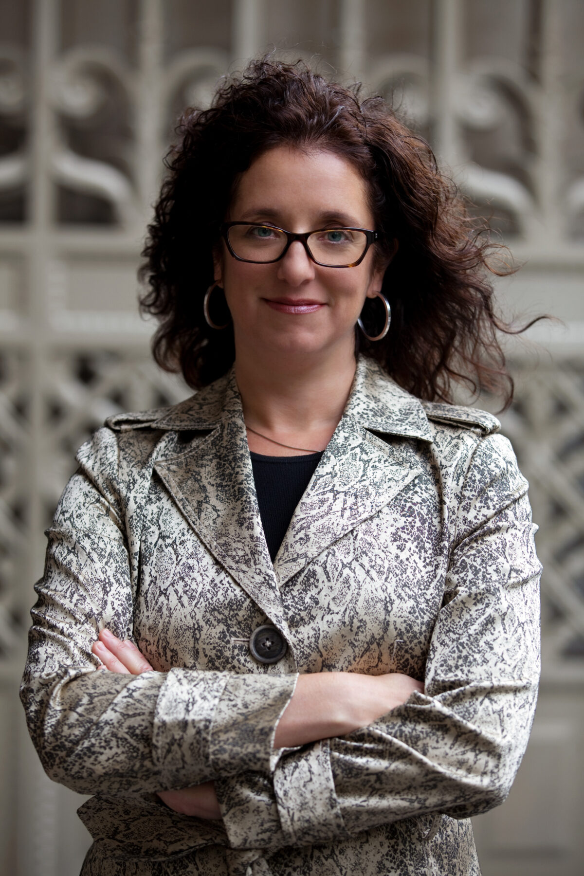 Headshot of a woman with her arms crossed looking at the camera. Her brown curly hair blows in the wind, and she is wearing a snake print silver blazer.