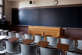 Image of an empty classroom. Chairs and desks face an empty chalkboard. A wooden desk stands in front of the chalkboard.