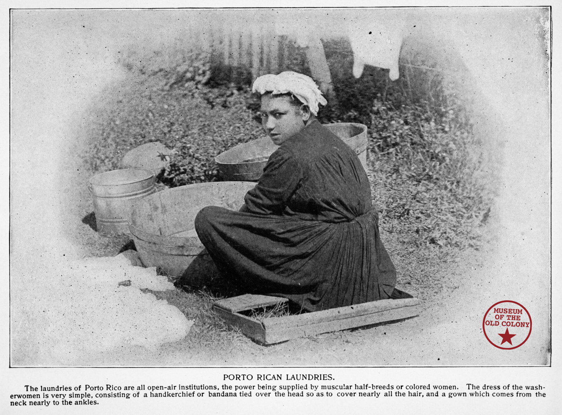A historic black and white photograph of a woman in a dark dress and light bonnet seated on the ground, looking behind her at the camera. The caption reads: "Porto Rican Laundries."