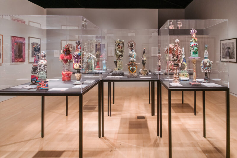 Installation view of Oletha DeVane: Spectrum of Light and Spirit. Various colorful sculptures are displayed in display cases at the center of a gallery.