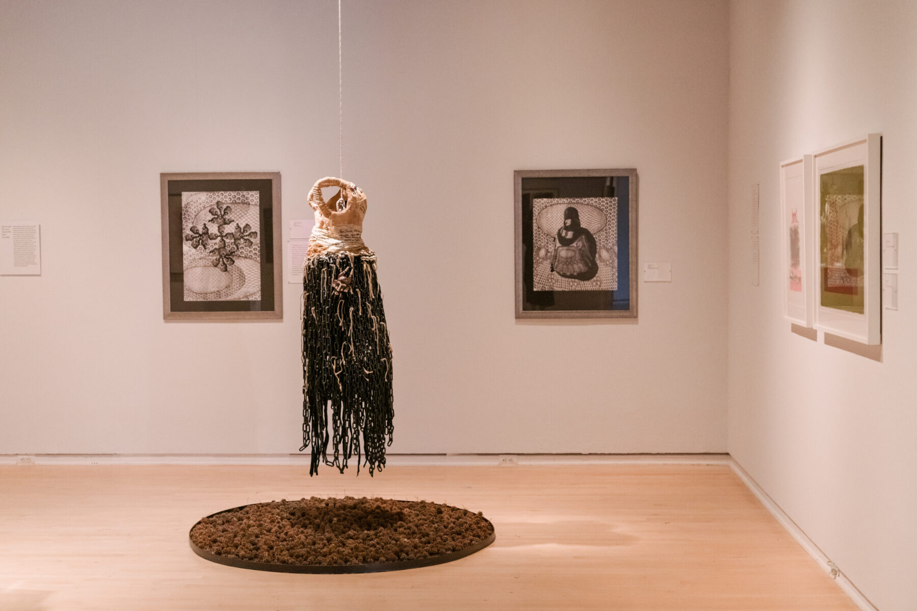 Installation view of Oletha DeVane: Spectrum of Light and Spirit. A dress-like sculpture hangs from the ceiling above a circular work on the wooden floor. Various framed works surround the installation on white walls.