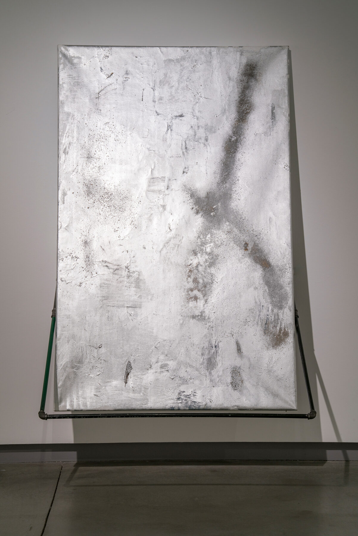 A silver metallic canvas leans against a white wall on a pedestal. A sight shadow is angled within the piece itself.