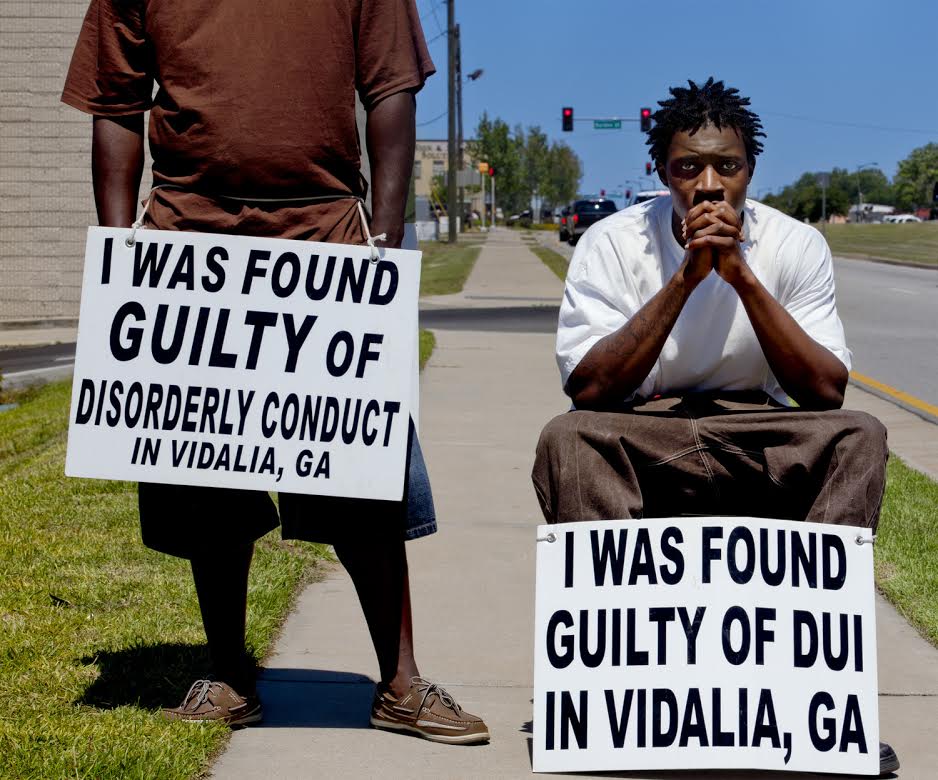 A photograph of two dark skinned men on a city sidewalk. A man, standing and wearing a maroon shirt, holds a sign that says "I was found guilty of disorderly conduct in Vidalia, GA." To his left, a man crouches and stares at the camera. He interlaces his hands and holds them near his face. He is wearing a white t-shirt and khaki shorts. His sign reads, "I was found guilty of DUI in Vidalia, GA."