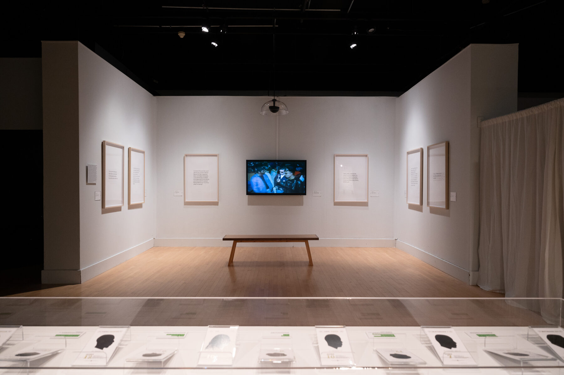 Installation view of Tahir Temphill: Rap Research Lab. A video is presented on a large TV screen with a wooden bench in front. In the foreground of the photograph is a glass display case filled with silhouettes.