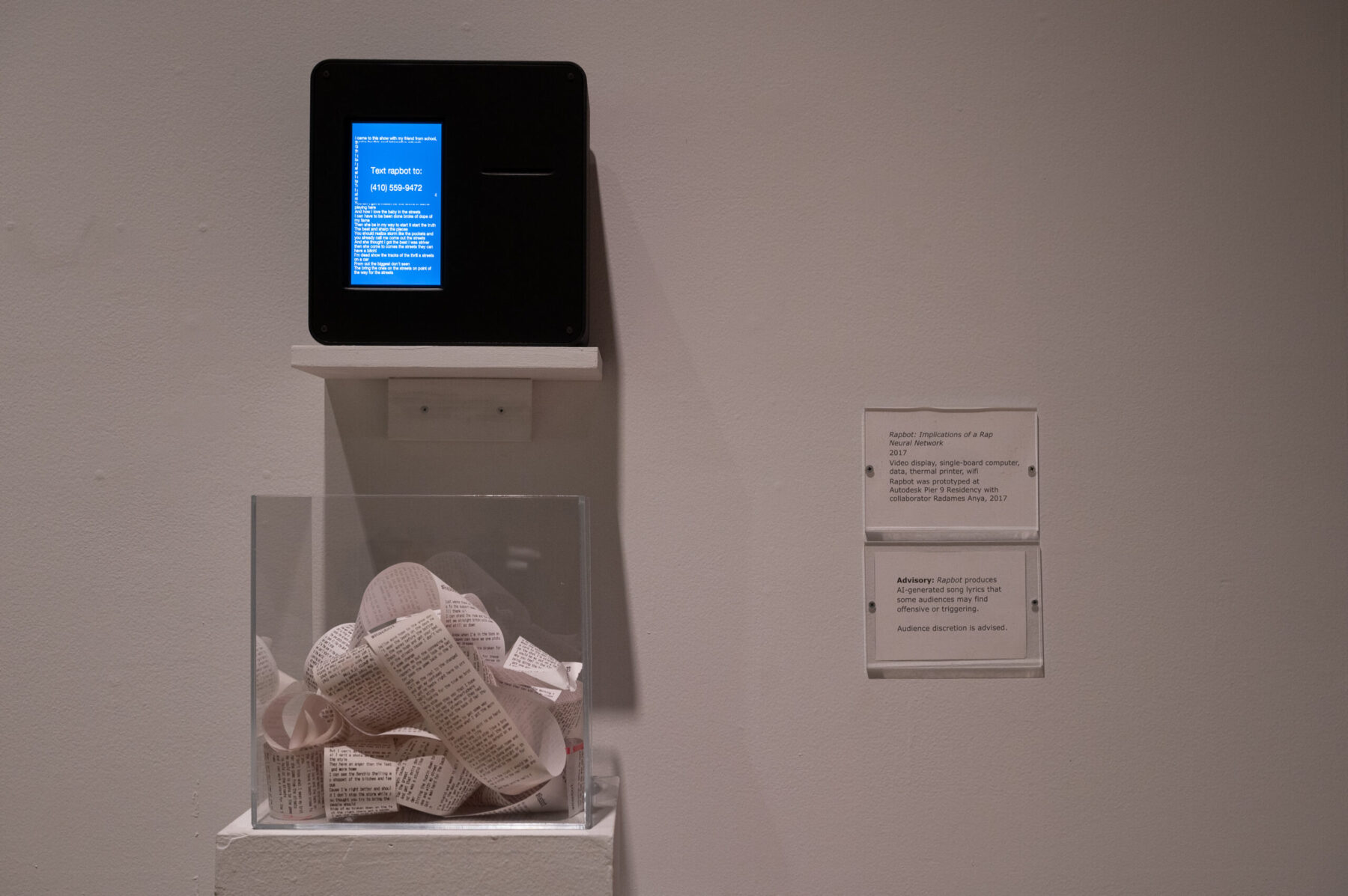 Installation of multimedia work by Tahir Temphill in Tahir Temphill: Rap Research Lab. The work consists of a screen and a glass box filled with a typed text on a long strip of paper.