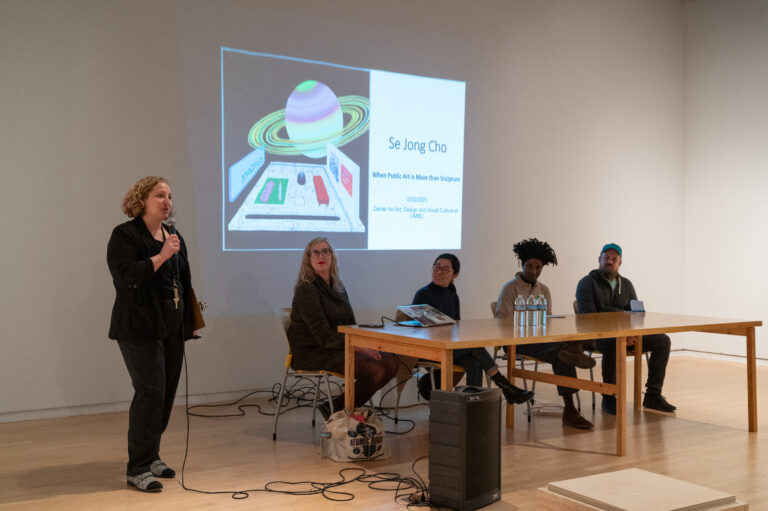 A woman with a black shirt and black pants holds a microphone and speaks towards an unseen audience. To her left are four panelists sitting at a light wood table. Behind the panel is a projection from a powerpoint slide about Se Jong Cho.