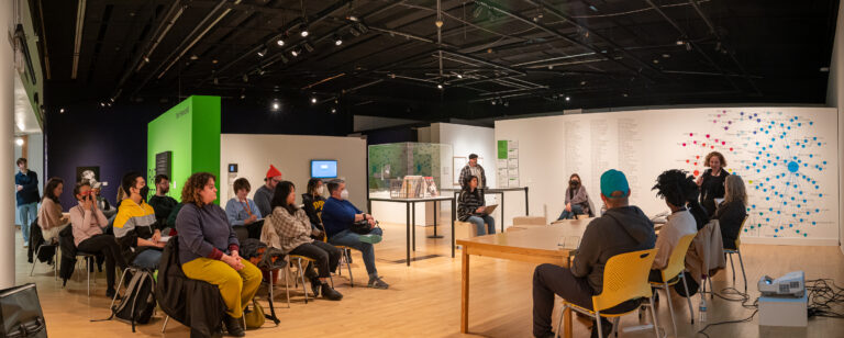 Panoramic view of CADVC's gallery space, in which a discussion panel is taking place. An audience is seated at left while panel members are seated in front of a long table at right. All are facing a woman standing behind the panel with a dark shirt and short blonde hair.
