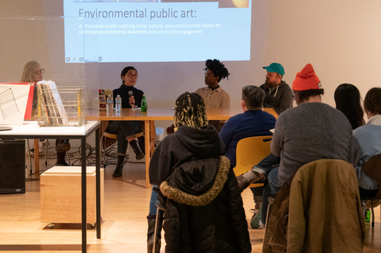 Scene of a indoor discussion panel. Five people are seated facing four individuals, speaking to each other. Projected behind them is a powerpoint slide for environmental public art.