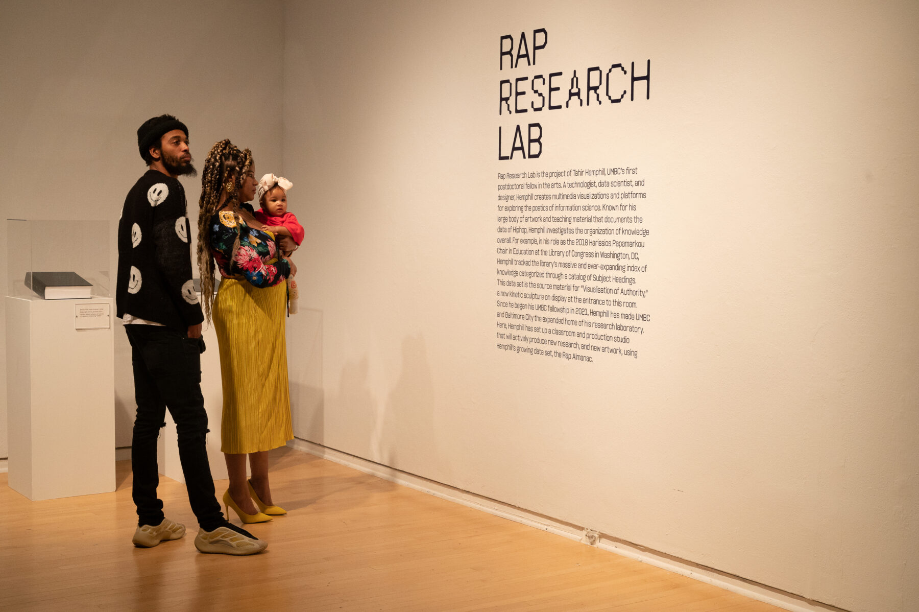 A man stands behind a woman who is holding a baby. The family is reading didactic label for "Rap Research Lab" exhibition.