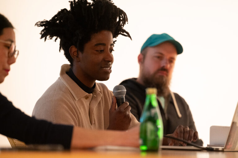 A woman with brown dreadlocks reads from a laptop while holding a microphone. She wears a taupe sweater with a collar. In the background is a bearded man wearing a dark hoodie and a teal colored baseball cap.
