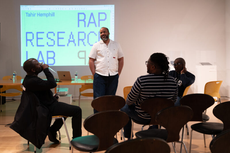 Tahir Temphill leading Rap Research Lab event. He stands with his hands in his pocket, wearing a short-sleeved white button-down shirt.
