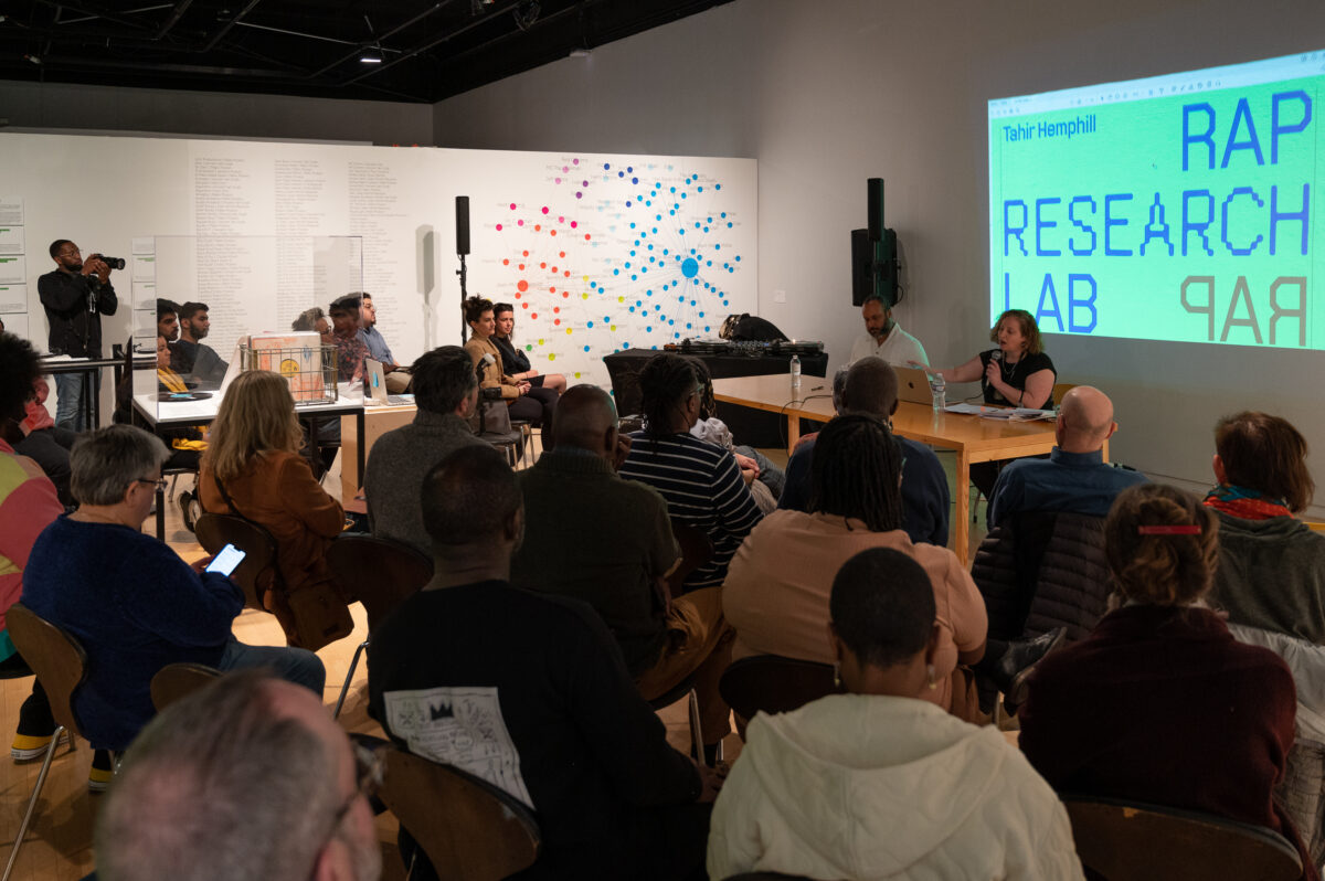 Room filled with people seated in front of Tahir Temphill and Rebecca Uchill. A teal-colored projection display reads "Rap Research Lab."