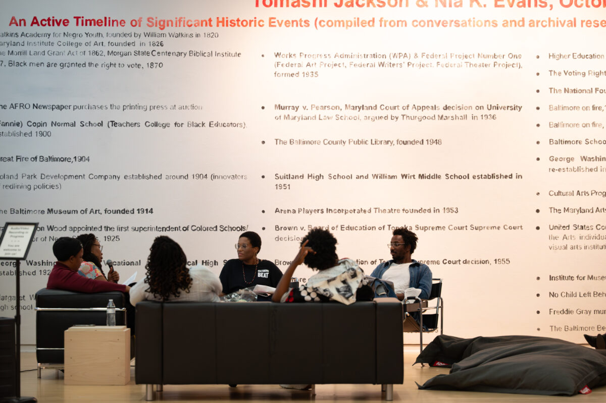 A group of people are seated on couches and chairs in front of a larger mural, which includes text about the exhibition, "Pedagogy Study Hall."
