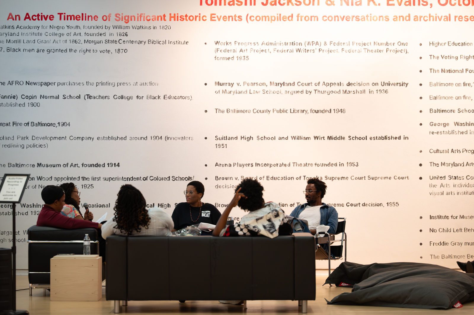 A group of people are seated on couches and chairs in front of a larger mural, which includes text about the exhibition, "Pedagogy Study Hall."