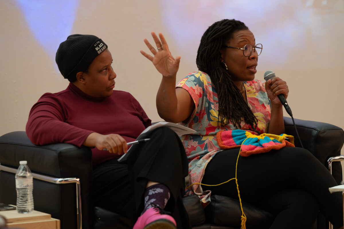 Tomashi Jackon, on the right hold a mic and gestures with her right hand toward Nia K. Evans seated beside her sitting cross-legged wearing a cap