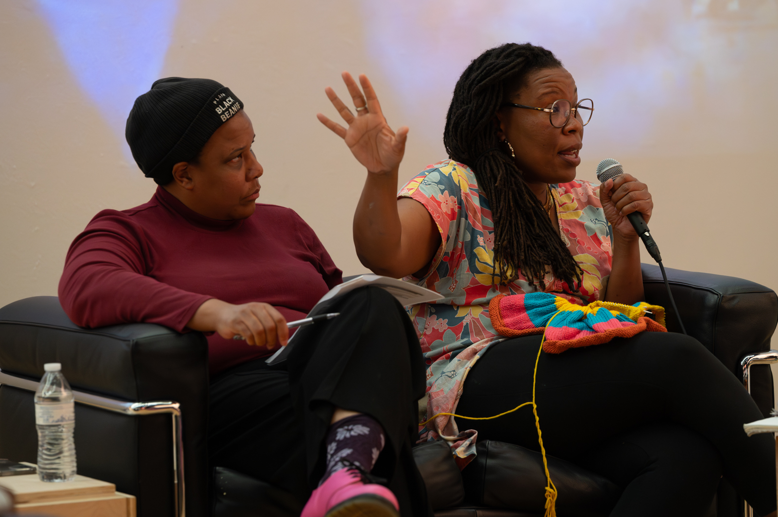 Tomashi Jackon, on the right hold a mic and gestures with her right hand toward Nia K. Evans seated beside her sitting cross-legged wearing a cap