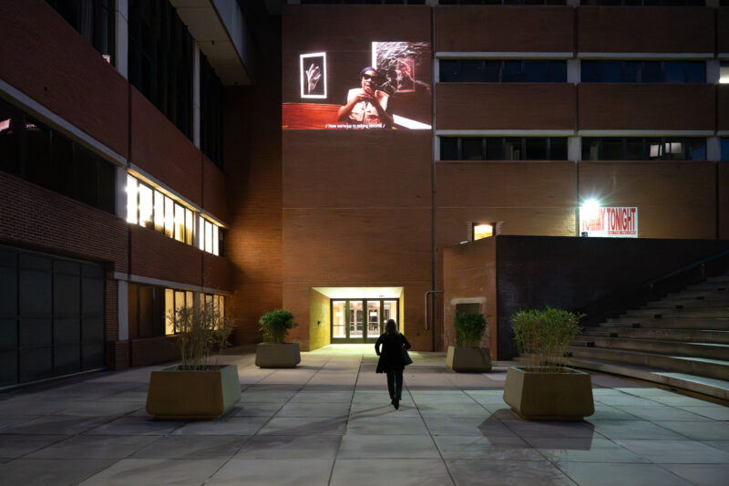 A nighttime exterior shot of a building with red brick. A video installation is projected on a wall with a person in shadow walking towards a doorway beneath it.