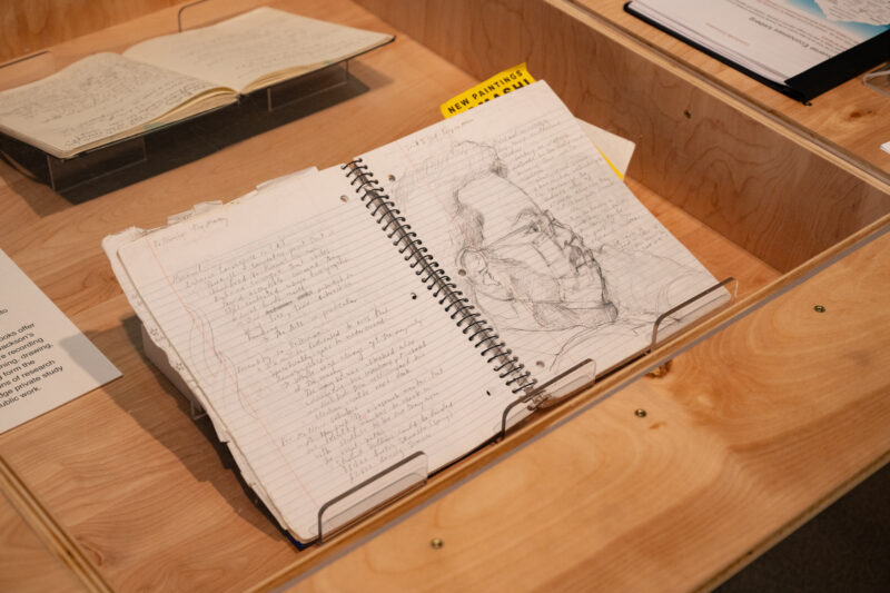 Open notebook featuring sketches and writing of an artist, placed in a wooden display case.