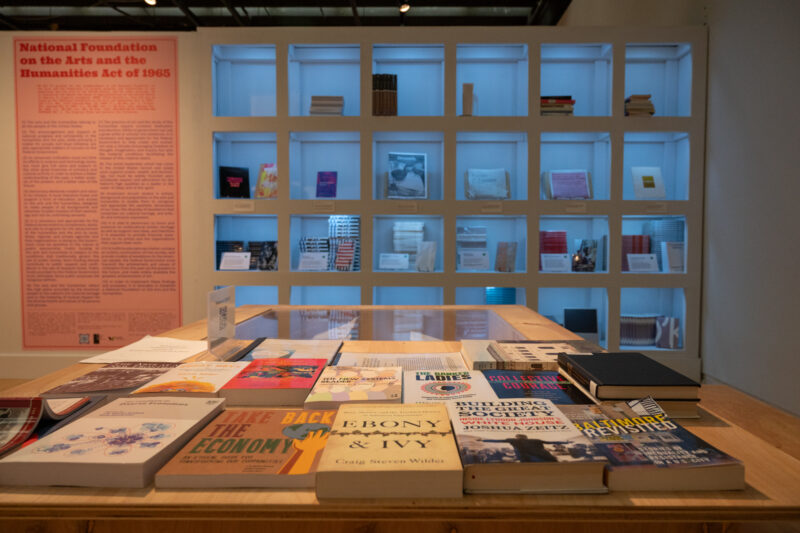 Selection of books stacked upon a wooden table with square shelves behind it.