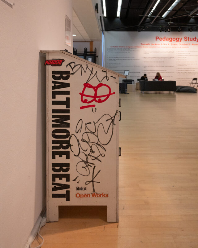 A graffitied newspaper stand for the Baltimore Beat installed in a white-walled gallery.