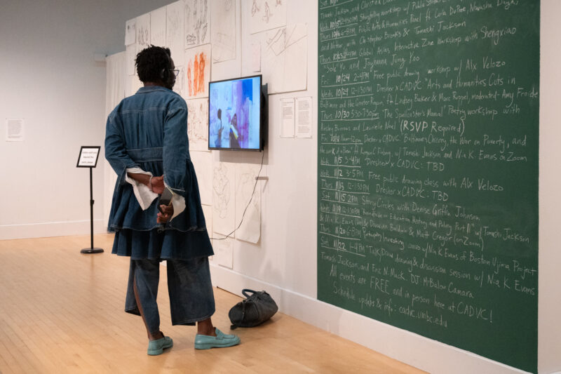 An individual with a dark skin-tone stands with his hands behind his back, watching a work of video art on a mounted TV screen. They wear a denim ensemble with light blue loafers. Next to them is green chalkboard with events written out in white chalk.