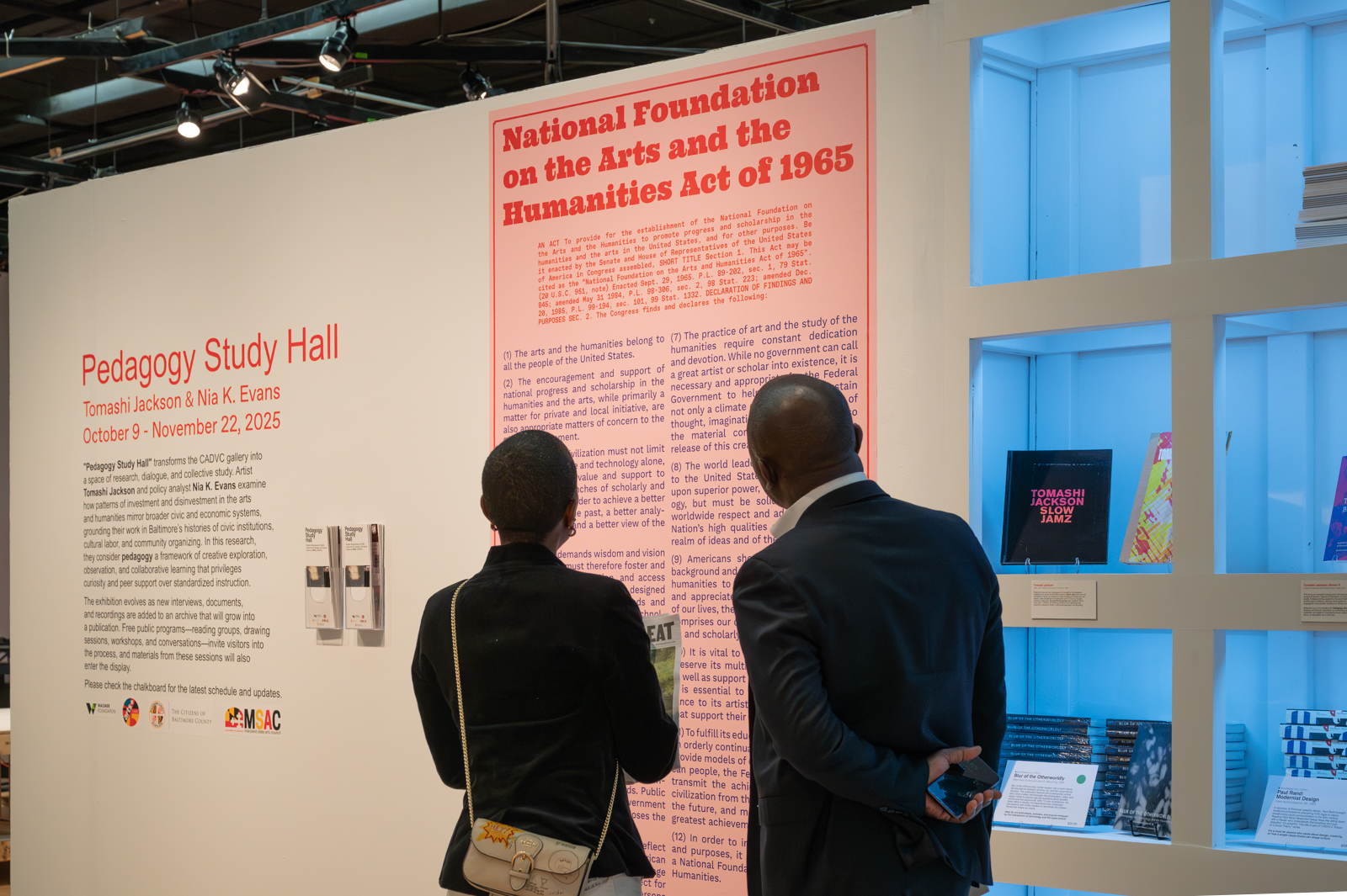 Two individuals with their back to the viewer stand side-by-side reading didactic exhibition text on a gallery wall. The panel is pink with red and black lettering.