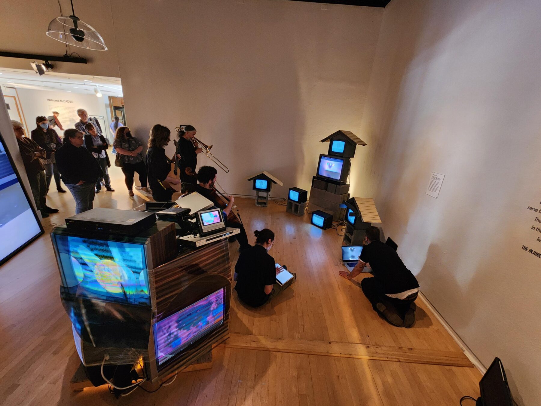 Various television monitors are arranged throughout a gallery space, both on pedestals or underneath makeshift roofs. Two people are seated on the floor, working on computers, which three other musicians play the trombone, guitar, and cello. All of them wear black.