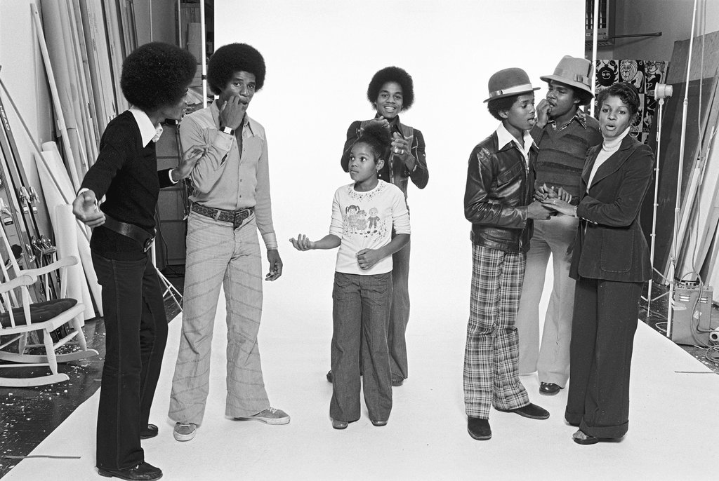 A candid black-and-white photograph of a Black family preparing for a photoshoot. They all wear attire popular in the 1970s, including bell-bottom pants and wide lapel shirts. Behind them is an illuminated white backdrop and other photograph props are seen to the left and right of the photo.