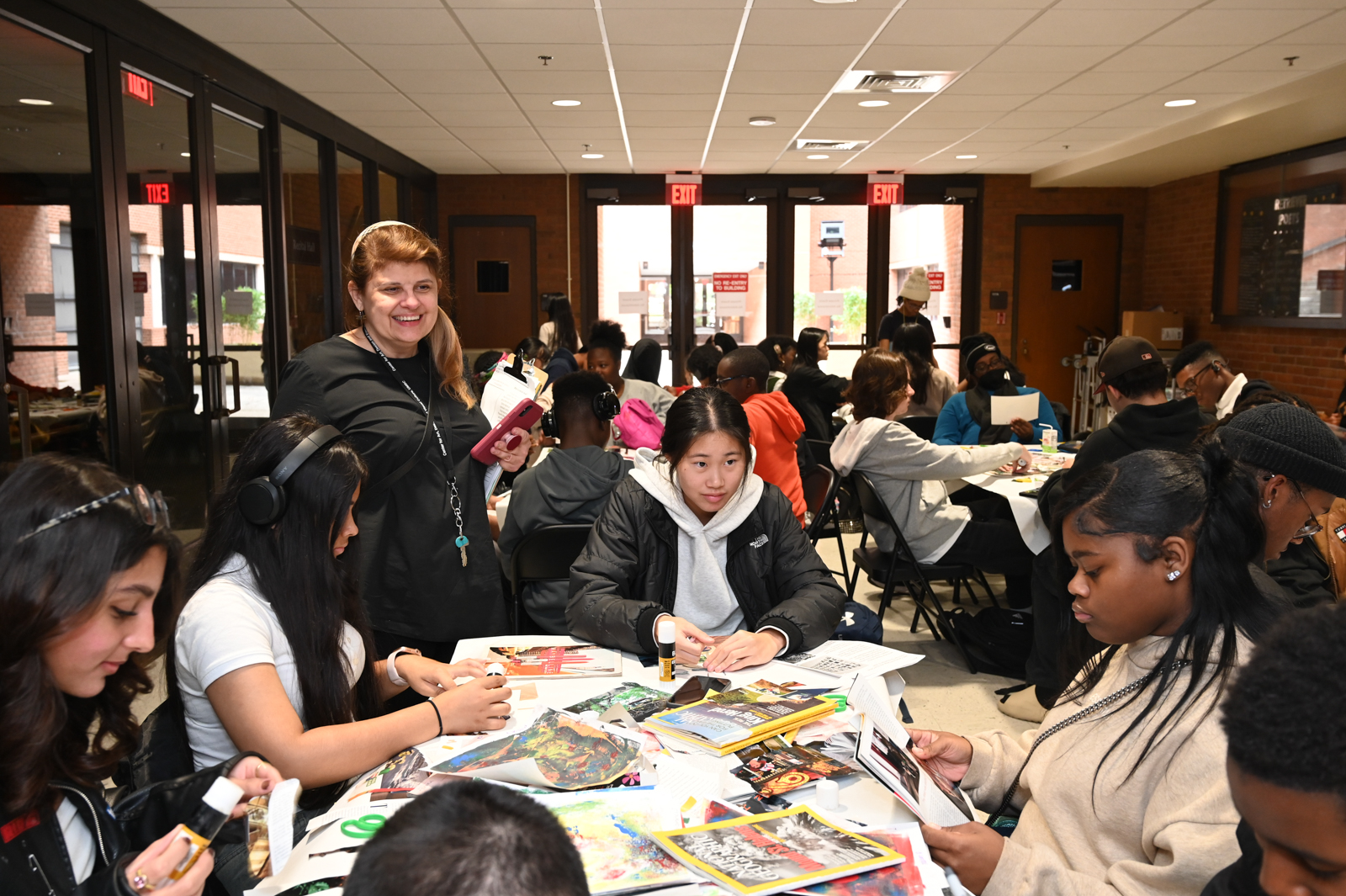 A group of teenagers are seated at a table, cutting and gluing pictures from magazines for a collage. A woman with a light skin-tone stands to their left. She wears a black outfit, a white headband, and a lanyard with a key.