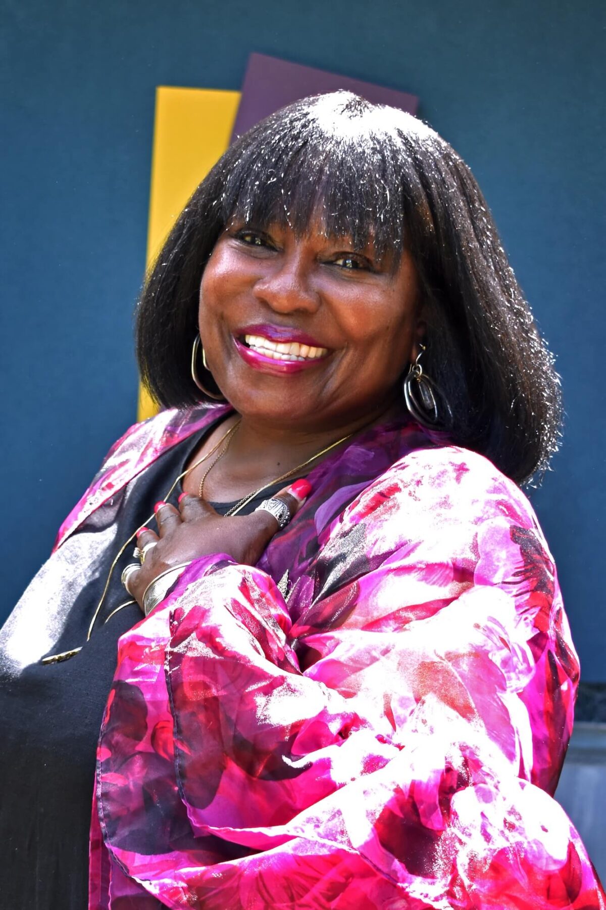 A portrait of a woman with a dark skin-tone smiling at the camera. She has a black bob and wears a bright fuchsia blouse.