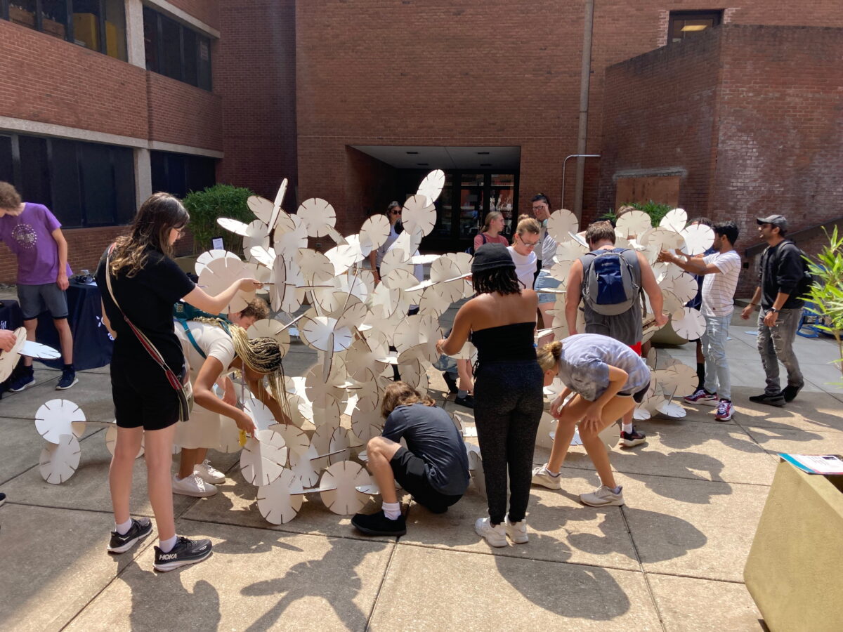 Various individuals stand around around standing sculptures of white interlocking plates. They are working together in an outdoor space with concrete beneath their feet.