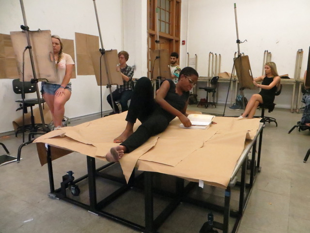 A woman with a dark skin-tone and a shaved hairstyle lays on her side upon a table covered in craft paper. Around her, students draw her figure on easels before them.