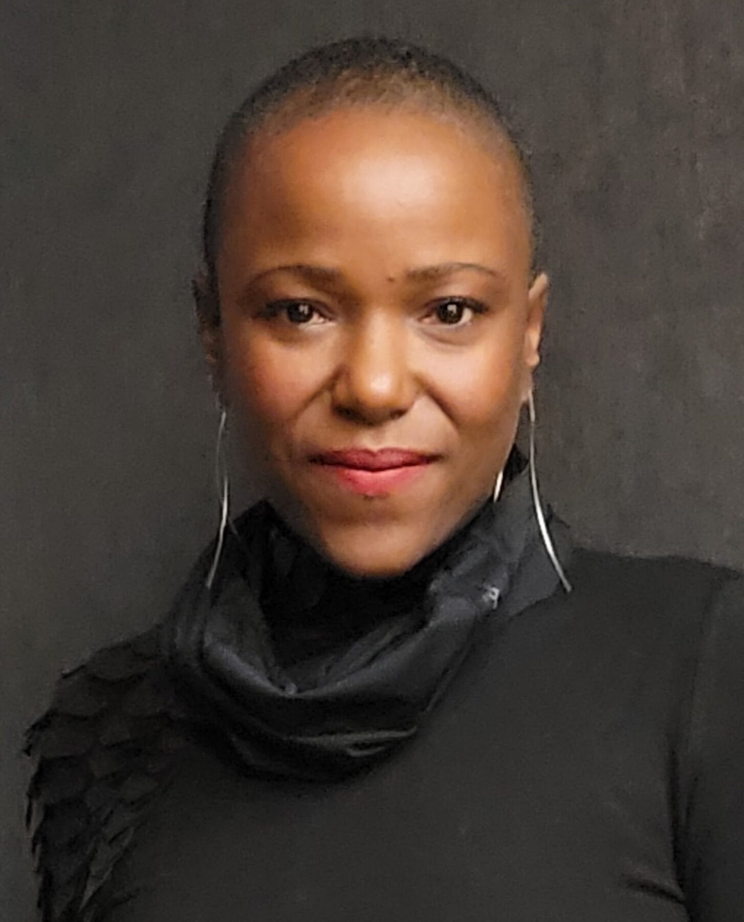 A portrait of a woman with a dark skin-tone with a shaved hairstyle who is wearing a black turtleneck with chandelier earrings.