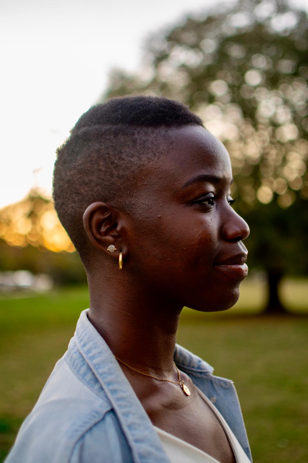 A portrait of a woman with a dark skin-tone and a shaved hairstyle. She wears silver earrings and a chambray shirt.