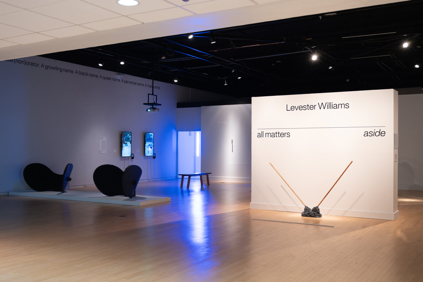 An installation view of a gallery space. On a central wall, "Levester Williams" and "all matters aside" are shown in black lettering. Below it is a mixed media sculpture with two wooden poles pointed in opposite directions from a metallic base.