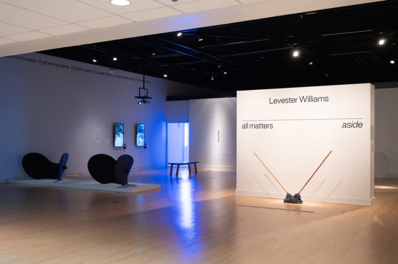 An installation view of a gallery space. On a central wall, "Levester Williams" and "all matters aside" are shown in black lettering. Below it is a mixed media sculpture with two wooden poles pointed in opposite directions from a metallic base.