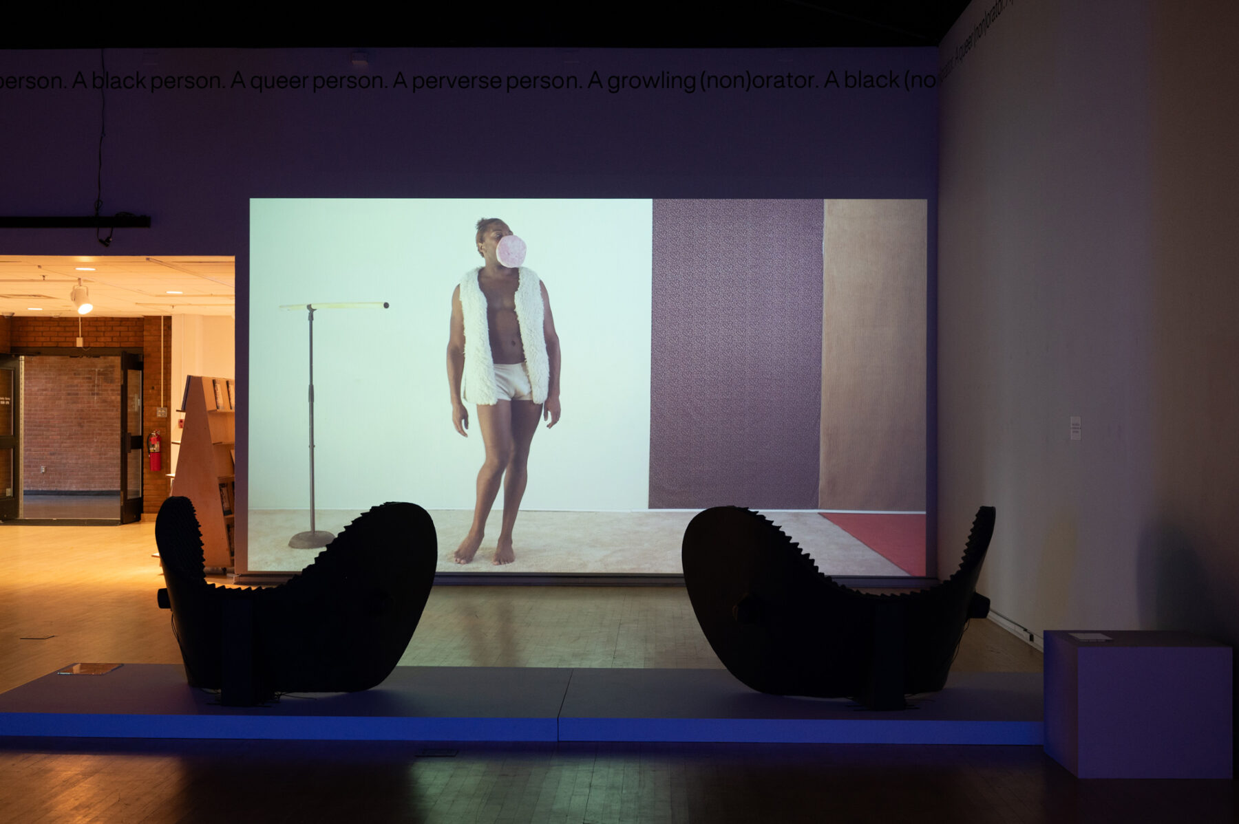 An installation view of a video projection. An individual with a dark skin-tone stands to the left side of the projection, wearing a white fur vest and white shorts. They are blowing a large pink bubblegum bubble. The rest of the scene is empty except for a black and khaki green panel to the subject's left.