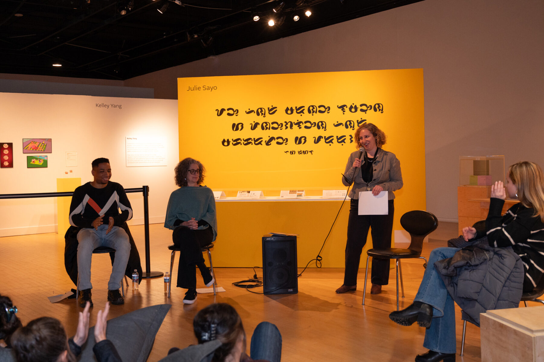 A man and a woman are seated to the right of another woman, who holds a microphone while talking to a larger audience. She has short blonde hair and wears an all black outfit aside from a khaki button-down shirt. Behind her is a yellow wall with scripted text.