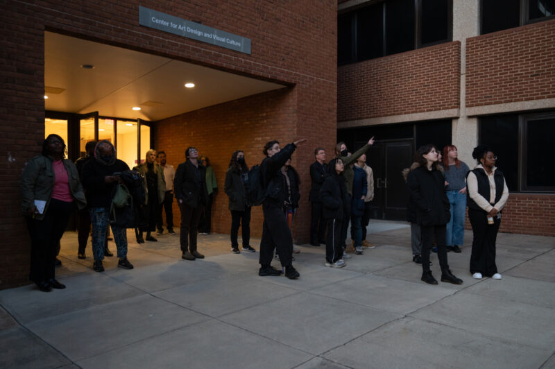 A large group of people stand outside a building entrance and look to the right side of the image. Towards the center of the group, two people point in the same direction everyone is looking.
