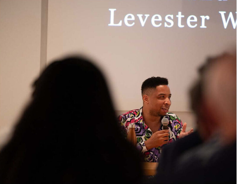 A man with a dark skin-tone and a colorful button-down shirt speaks to an audience through a microphone, which he holds in his right hand.