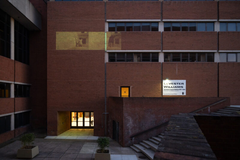 An exterior view of a red brick building. A yellow-toned video is projected between the second and third floors of the building. At the base of a the building near a set of outdoor stairs is a white banner that reads "Levester Williams" black text.