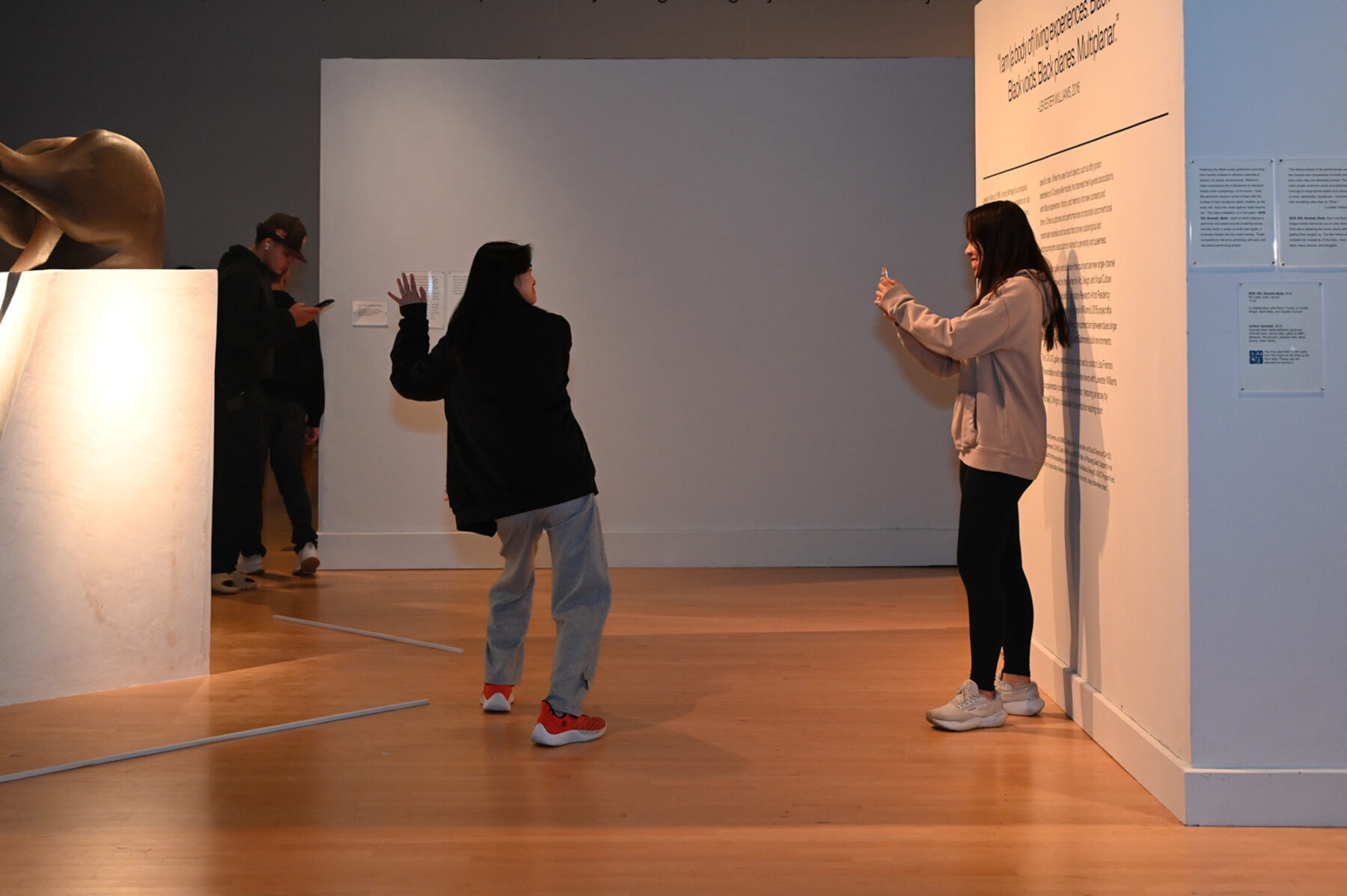Two women stand in a gallery space. The women at left wears black jacket, grey pants, and red sneakers. She is posing with her arms up. To the right is a woman in a grey hoodie, black leggings, and white sneakers. She holds up the camera to take a picture of the other, smiling while doing so.
