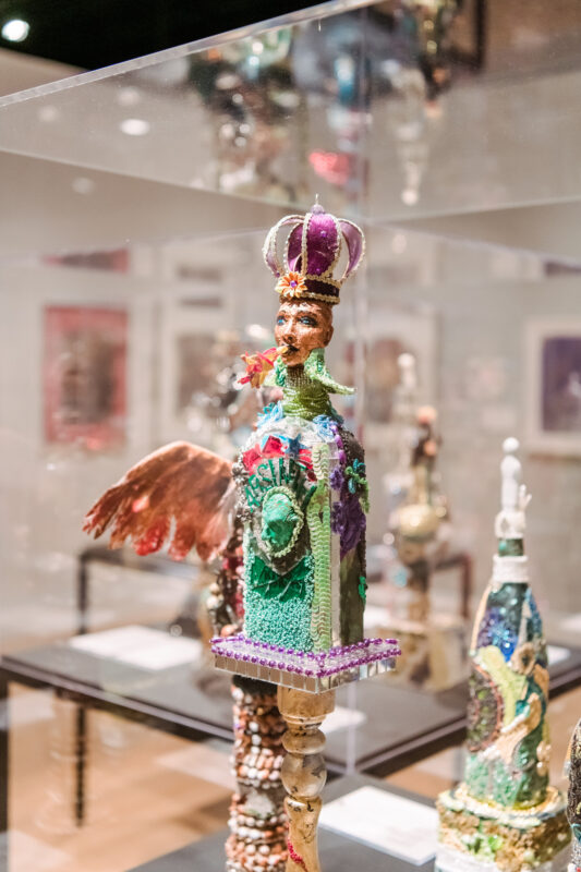 Detail of a small sculptural bust, showing a person wearing a purple crown with a predominantly green ensemble. The sculpture is placed upon an ornate beaded pedestal.