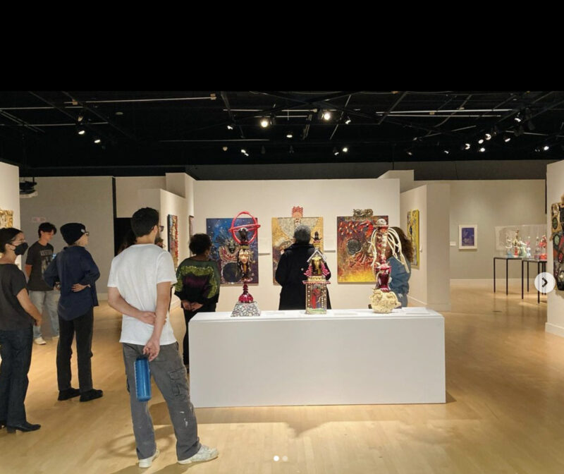 Individuals mingle about a gallery space, looking at multimedia artwork on a pedestal or on the walls around them.